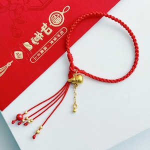 May include: Red braided bracelet with a gold-colored gourd charm and tassel accents. The bracelet is displayed on a red card with gold Chinese characters and illustrations. The bracelet is a symbol of good luck.