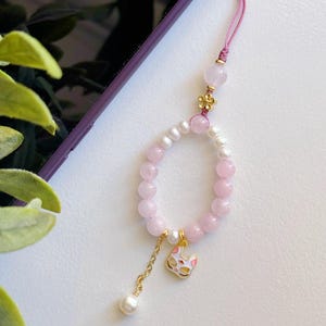 May include: A pink and white beaded phone charm with a gold cat charm and a pink flower charm. The charm is attached to a pink string.