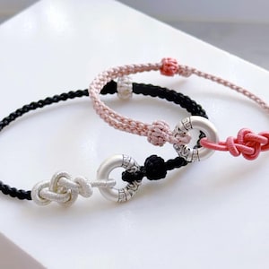 Peut inclure: Deux bracelets faits à la main. L'un est noir, l'autre rose. Chaque bracelet présente un motif de cordon tressé et un nœud décoratif. Les bracelets sont reliés par un anneau argenté avec un motif gravé.