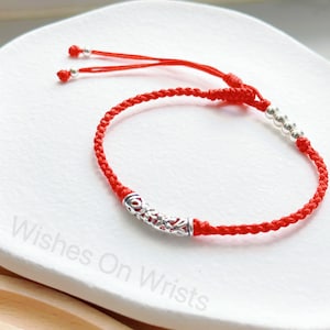 May include: Red braided bracelet with silver-colored accents. The bracelet features a decorative silver tube and several small silver beads. The adjustable closure has two small silver beads. The bracelet is displayed on a white surface.