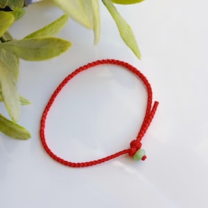 Peut inclure: Un bracelet en cordon rouge avec une perle verte. Le bracelet est noué et a un pompon.