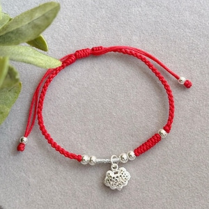 Puede incluir: Una pulsera trenzada roja con un dije plateado. La pulsera presenta cuentas plateadas y un dije plateado decorativo. La pulsera está hecha de un cordón rojo y tiene extremos ajustables.