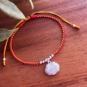 May include: A red and gold braided string bracelet with a silver charm. The charm is a round, ornate design with a raised center.