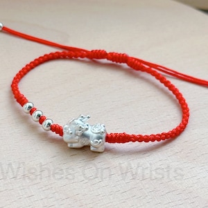May include: A red string bracelet with a silver charm in the shape of a mythical creature. The bracelet is tied with a knot and has silver beads.