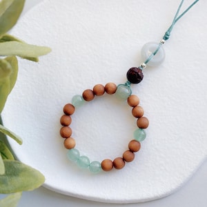 May include: A wooden and green stone beaded bracelet with a jade pendant and a brown wooden bead attached to a teal cord.