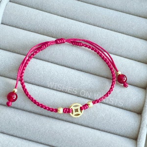 May include: A magenta-colored bracelet with a woven cord design. The bracelet features a gold-colored charm with a square design, gold beads, and red beads. The text "WISHES ON WRISTS" is visible in the background.