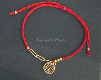May include: Red string bracelet with a gold charm featuring a Chinese character. The bracelet has gold beads and a gold chain link.