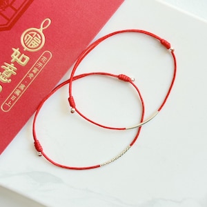 Könnte beinhalten: Zwei rote Kordelarmbänder mit verstellbaren Schiebeknoten und silberfarbenen Metallakzenten. Ein Armband verfügt über einen glatten, gebogenen Metallstab, während das andere ein strukturiertes, funkelndes Metallsegment aufweist.