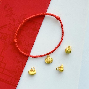 May include: A red braided bracelet with a gold charm and three gold charms scattered around it. The bracelet has a circular design with a gold charm that has Chinese characters on it. The other charms are in the shape of an apple, a cloud, and a gourd.