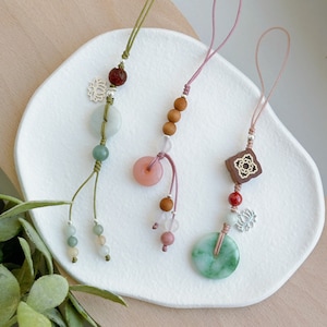 Puede incluir: Tres colgantes para teléfono con cuentas y dijes de diferentes colores. Los dijes incluyen un círculo verde jade, un círculo rosa y un círculo verde jade con un dije de flor de loto plateado. Los dijes están unidos a cordones verdes, rosas y marrones.