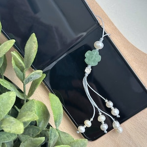 Op de afbeelding: Een groene bloemenbedel met een zilveren kraal en witte parel accenten hangt aan een grijze kwast. De bedel is bevestigd aan een telefoonhoesje.
