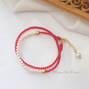 May include: Red string bracelet with white pearls and gold beads. The bracelet has a gold clasp and a small pearl charm. The text "Wishes On Wrists" is visible in the image.
