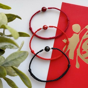 May include: Three red string bracelets with black and red beads. The bracelets are stacked on top of each other and are on a red background.