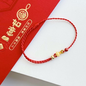 Puede incluir: Una pulsera de hilo rojo con detalles dorados, que incluye un pequeño dije y cuentas. La pulsera se muestra sobre un fondo rojo con caracteres chinos dorados.