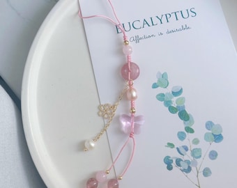 May include: A pink and gold phone charm with a butterfly, flower, and pink and white beads. The charm is hanging from a pink string. The text "EUCALYPTUS Affection is desirable." is visible in the background.