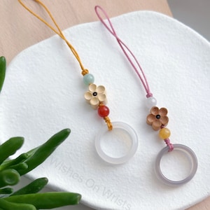May include: Two phone charms with a white and gray ring, a flower charm, and beads. The charms are on a white plate with the text "Wishes On Wrist" on the bottom.