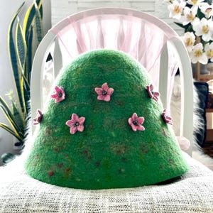 May include: A dome-shaped green felted hat featuring several small, five-petalled pink flowers attached to the exterior. Each pink flower has a dark, textured centre. The green felt material has a mottled, variegated appearance with subtle darker tones.