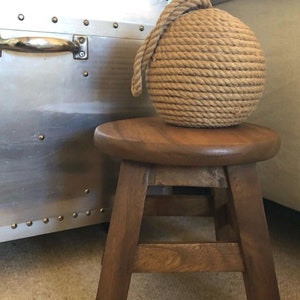 May include: A brown wooden stool with a rounded top and three legs. A natural rope doorstop sits on top of the stool.