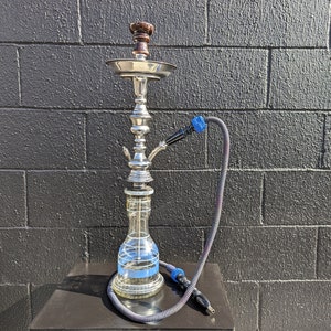 May include: A silver hookah with a clear glass base and a blue hose. The hookah has a brown wooden bowl and a silver metal stem.