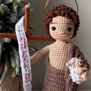 May include: Crocheted amigurumi doll of a shepherd holding a white lamb. The doll is wearing a brown robe and has a halo above its head. The doll is standing next to a wooden stick with a white ribbon that has red text on it.