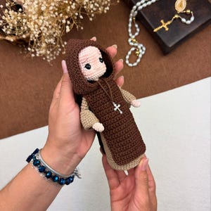 Pode incluir: Uma boneca de crochê vestindo uma túnica marrom com capuz. A boneca tem um rosto bege com olhos pintados e uma cruz de prata pendurada na túnica.