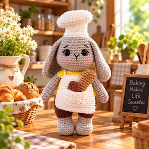 May include: A grey crocheted rabbit doll wearing a white chef hat and a white apron over a yellow shirt. The apron pocket contains a brown cookie and a tan breadstick. The rabbit has black eyes, pink cheeks, and brown shorts.