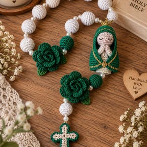 Op de afbeelding: Een handgemaakte gehaakte rozenkrans met witte bolvormige kralen, twee donkergroene gehaakte rozen en een centrale figuur van een gekroond persoon in een groene mantel met gevouwen handen. De rozenkrans eindigt met een groen en wit gehaakt kruis.