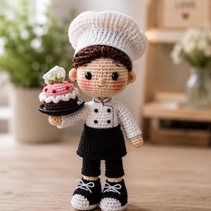 May include: A crochet doll dressed as a chef, wearing a white toque, a white long-sleeved jacket with black buttons, a black apron, black trousers, and black trainers with white laces. The doll holds a small black plate with a pink cupcake.