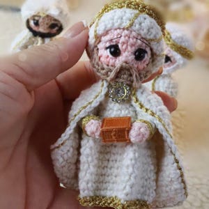 Könnte beinhalten: Gehäkelte Figur eines Weisen, der ein weißes Gewand mit goldenen Verzierungen und eine passende Kopfbedeckung trägt. Die Figur hat einen braunen Bart und hält eine kleine orangefarbene Truhe. Ein funkelnder Schmuck ziert die Brust. Weitere ähnliche Figuren sind im Hintergrund.