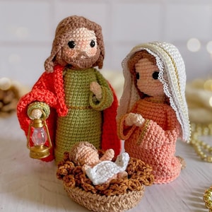 May include: A crochet nativity set featuring three figures. One figure has brown hair and a beard, wearing a green tunic and a red cloak, holding a small gold lantern. Another figure wears a peach dress and a white lace-trimmed head covering. A small infant lies in a brown crochet manger.