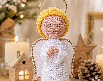 May include: A crocheted angel figurine with a pale pink head, yellow hair, and a white dress. The angel has white wings with gold trim and a gold halo above its head. The hands are clasped together in front of the chest in a prayer position.