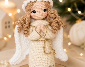 May include: A crocheted angel doll with light tan skin, curly blonde yarn hair, and a white halo featuring small white flowers. The doll wears a cream-colored dress with gold trim, a gold waist tie, and white wings, with hands clasped in a prayer position.