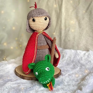 May include: A handmade crochet doll of a knight in gray armor, a red cape with gold trim, and a helmet. A green crochet dragon sits at the knight's feet. The doll is on a wooden slice base.