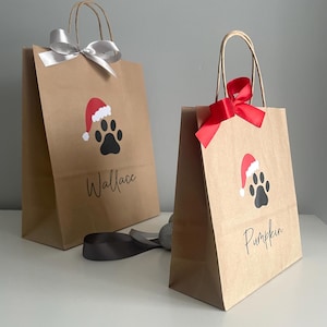 May include: Two brown paper gift bags with red and white Santa hats and black paw prints. The bags are decorated with black text that says "Wallace" and "Pumpkin".