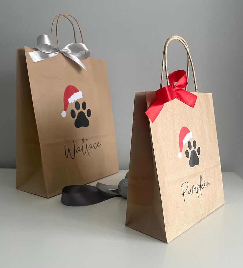 May include: Two brown paper gift bags with red and white Santa hats and black paw prints. The bags are decorated with black text that says "Wallace" and "Pumpkin".