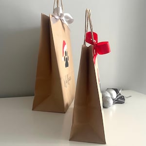 May include: Two brown paper gift bags with handles. The larger bag has a white ribbon and a paw print with a Santa hat on it. The smaller bag has a red ribbon. Both bags have the word "Woof" written on them.