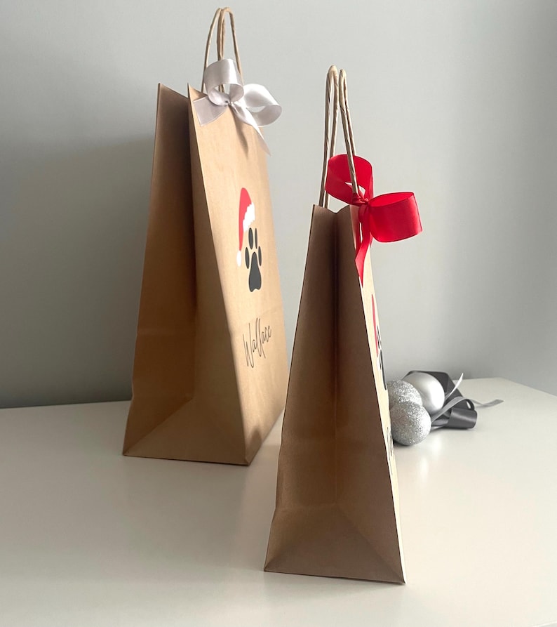 May include: Two brown paper gift bags with handles. The larger bag has a white ribbon and a paw print with a Santa hat on it. The smaller bag has a red ribbon. Both bags have the word "Woof" written on them.