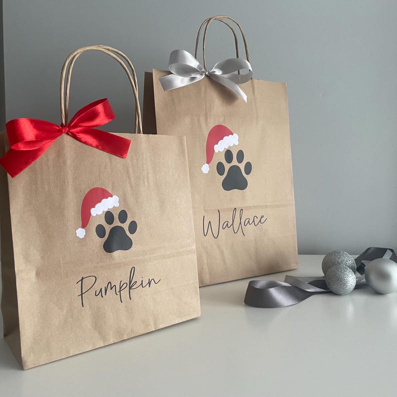 May include: Two brown paper gift bags with red and white Santa hats and paw prints. The bags are decorated with the names "Pumpkin" and "Wallace".