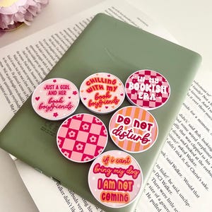 May include: Six circular phone grips with different designs and slogans, such as "Chilling with my Book Boyfriends" and "If I Can't Bring My Dog I Am Not Coming." The grips are in shades of pink, orange, and white.