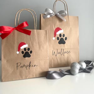 May include: Two brown paper gift bags with red and silver ribbon. The bags have a paw print with a Santa hat on them. The bags are personalized with the names "Pumpkin" and "Wallace".