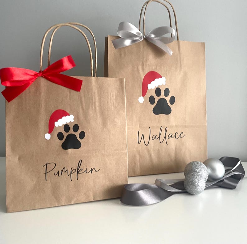 May include: Two brown paper gift bags with red and silver ribbon. The bags have a paw print with a Santa hat on them. The bags are personalized with the names "Pumpkin" and "Wallace".