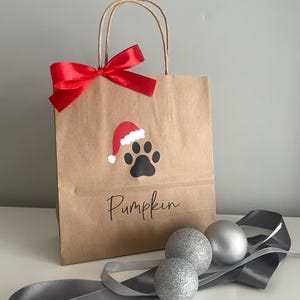May include: A brown paper gift bag with a red bow and a paw print with a Santa hat on it. The bag says "Pumpkin" on the front. There are three silver glitter ornaments and a gray ribbon on a white surface.