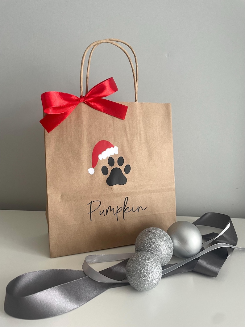 May include: A brown paper gift bag with a red bow and a paw print with a Santa hat on it. The bag says "Pumpkin" on the front. There are three silver glitter ornaments and a gray ribbon on a white surface.