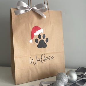 May include: A brown paper gift bag with a silver ribbon bow. The bag has a black paw print with a red Santa hat on top. The name "Wallace" is written in black below the paw print.
