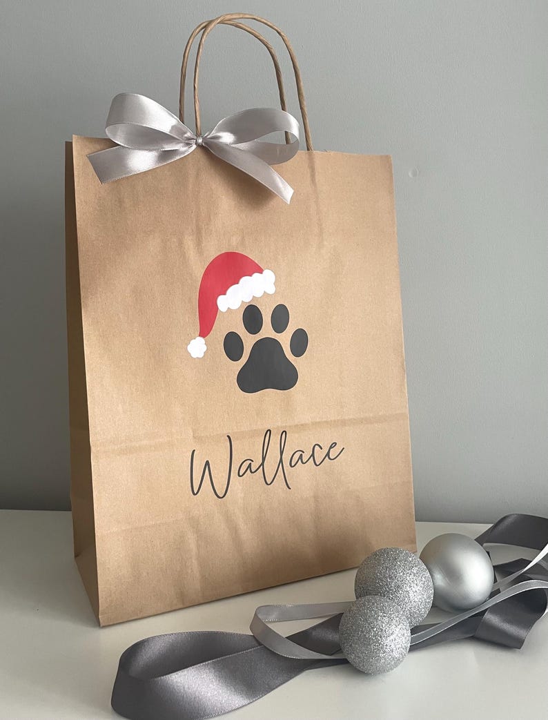 May include: A brown paper gift bag with a silver ribbon bow. The bag has a black paw print with a red Santa hat on top. The name "Wallace" is written in black below the paw print.