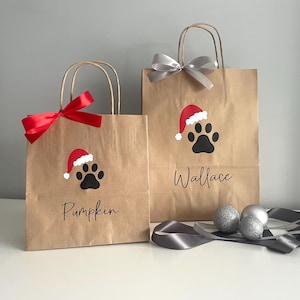 May include: Two brown paper gift bags with red and silver ribbon. The bags have a paw print with a Santa hat on them. One bag says "Pumpkin" and the other says "Wallace".