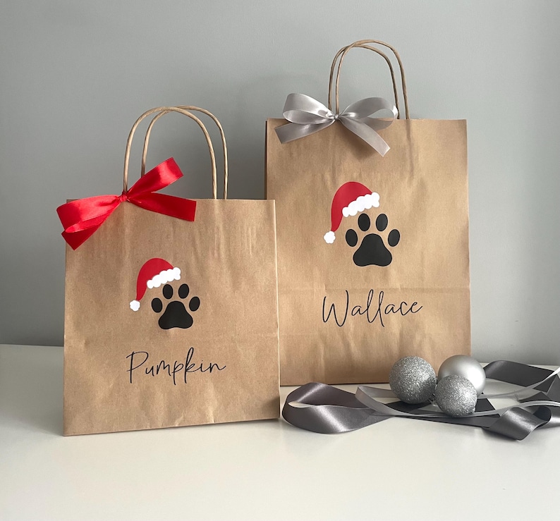 May include: Two brown paper gift bags with red and silver ribbon. The bags have a paw print with a Santa hat on them. One bag says "Pumpkin" and the other says "Wallace".