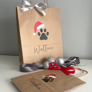 May include: Two brown paper gift bags with a paw print design and a Santa hat. The bags are personalized with the names "Wallace" and "Pumpkin".