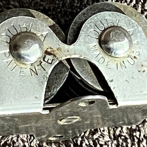 May include: A silver knife sharpener with a red handle. The sharpener has two sets of wheels with the text "DUBLIA PATENT" and "DUBLIA MADE IN U.S.A." on them.