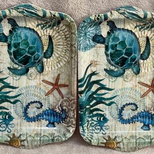 May include: Two rectangular trays with raised edges, featuring a nautical print of a teal sea turtle, a blue seahorse, a reddish-brown starfish, and green seaweed on a cream-colored background with faint map lines and circular patterns.